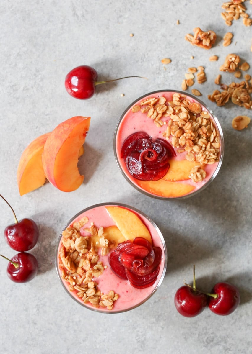 Two glasses of Peach-Cherry Smoothie and Coconut Chia Pudding Parfaits topped with a cherry rose and granola with cherries, peach slices and granola on the counter behind.