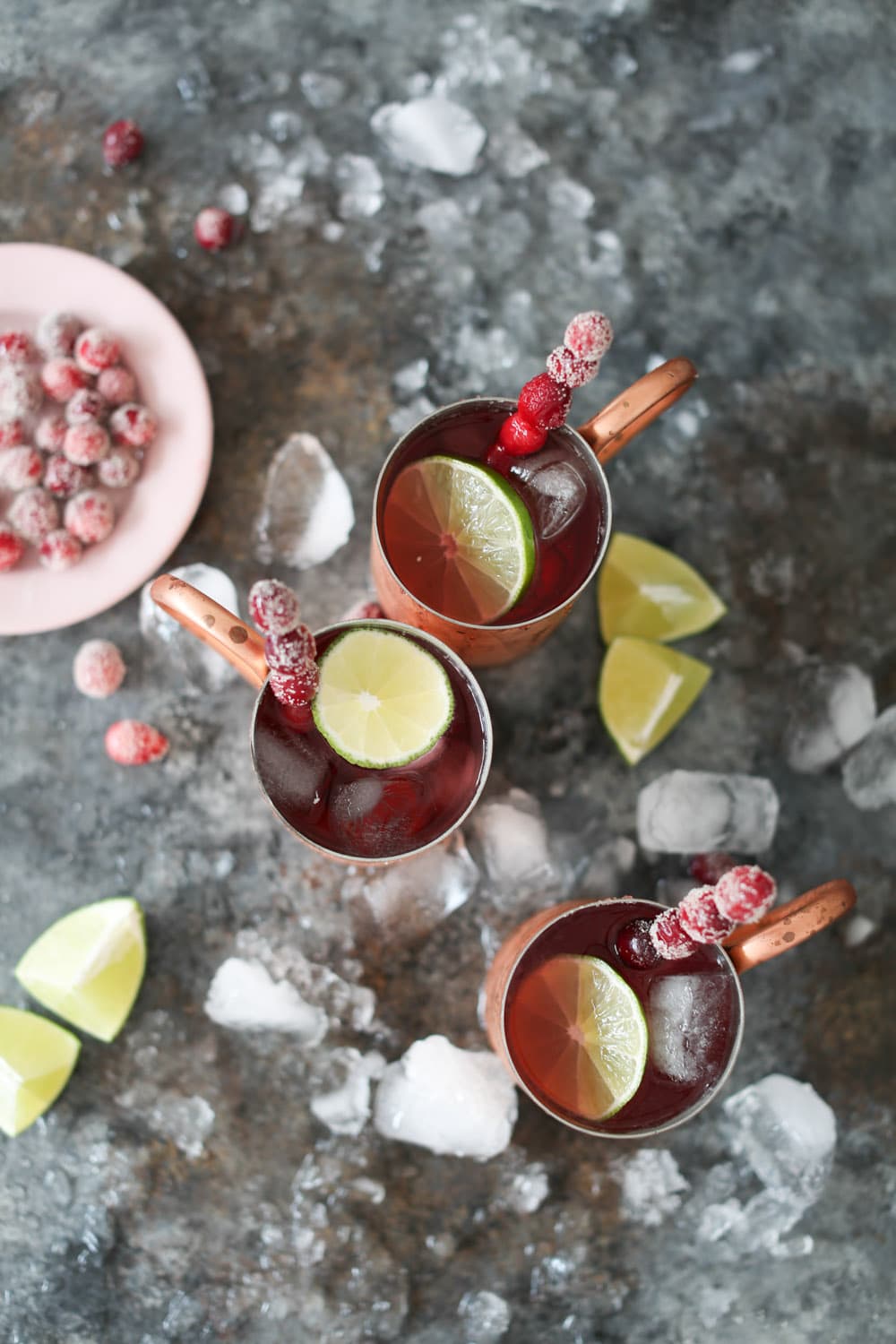 Overhead shot of three holiday-themed Moscow Mule cocktails with cranberry juice surrounded by crushed ice, lime wedges, and sugared cranberries.